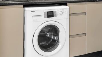 Un lave-linge BEKO à chargement frontal, de couleur blanche, se trouve dans une cuisine avec des armoires de couleur beige et un comptoir noir.