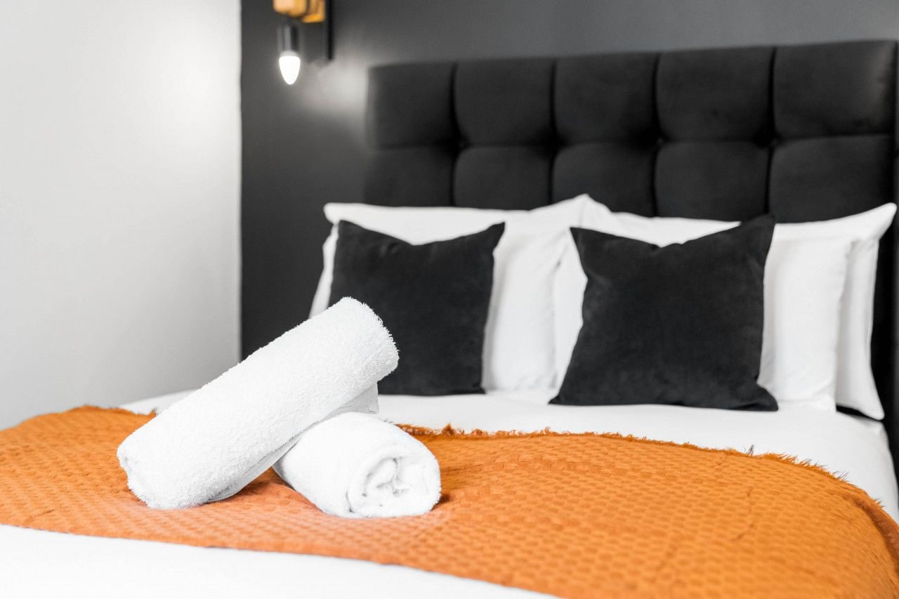 Une chambre d’hôtel avec une tête de lit capitonnée, des oreillers blancs et noirs, une couverture de lit orange à texture et une serviette blanche roulée.