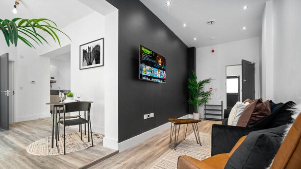 A modern living room with a black accent wall, a mounted TV, a brown leather sofa, and a dining area with a table and chairs.