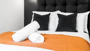 A neatly made bed with a white and orange bedspread, black and white pillows, and a rolled white towel.