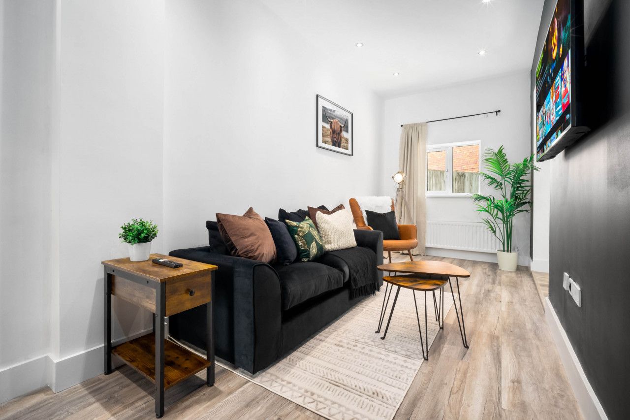 Un salon moderne avec un canapé noir, une table basse en bois, une table d’appoint avec une plante et un téléviseur à écran plat fixé au mur.