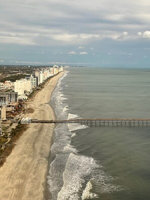 Vista aérea de una playa con un muelle que se adentra en el océano, junto a una hilera de edificios.