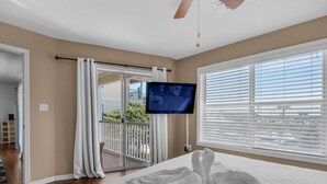A bedroom with a bed, a television, a balcony with curtains, and a view of the outside.