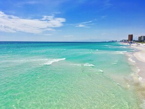 A beach with clear turquoise water, a sandy shore, and a city skyline in the background. - 5-min. walk to beach. Free parking. 4 Pools. Pickleball. King bed. Fenced yard. (Panama City Beach)