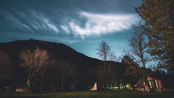A night sky with stars and clouds over a landscape with trees and a small building.