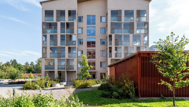 Un edificio de varios pisos con techo plano, ventanas grandes y un pequeño jardín con un árbol y arbustos.