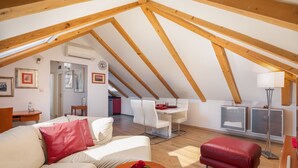 Un loft spacieux avec de hauts plafonds, des poutres apparentes, un coin repas, un salon avec un canapé et un pouf, et une petite cuisine.