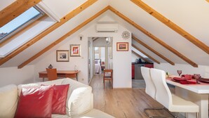 Une chambre confortable située dans les combles, avec un plafond en pente, des poutres en bois, un puits de lumière, un coin repas avec une table et des chaises, et une cuisine à l’arrière-plan.