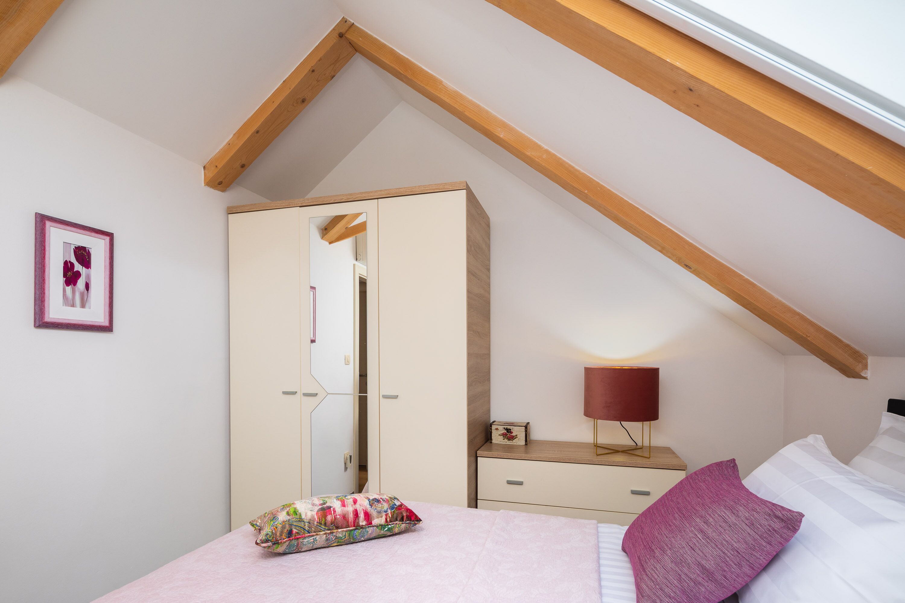A bedroom with a sloped ceiling, a bed with white and pink bedding, a wooden nightstand with a lamp, a wardrobe, and a framed picture on the wall.