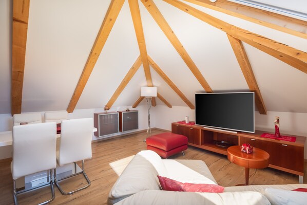 A modern loft bedroom with a sloped ceiling, wooden beams, a large flat-screen TV, a red ottoman, and a white sofa.