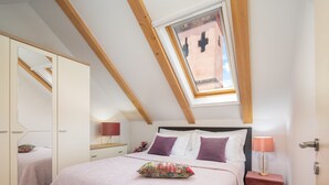 Une chambre avec un plafond en pente, un puits de lumière, un lit recouvert d’une couverture rose, une table de chevet avec une lampe et un miroir.