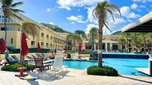 Poolområde på et resort med liggestole, palmer og en klar blå himmel.