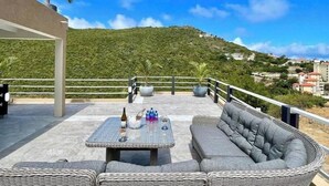 En tagterrasse med et bord dækket til to personer, en sofa og udsigt til en grøn bakkeside.