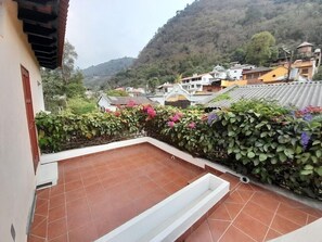 Eine Dachterrasse mit Terrakottafliesen, einem weißen Pflanzgefäß und einem üppigen, grünen Heckenstreifen mit rosa Blüten.