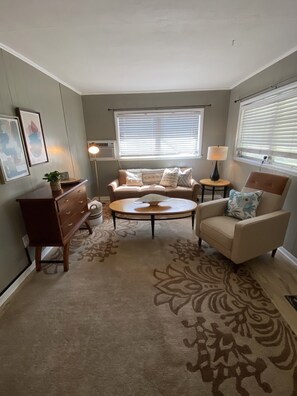 A living room with a sofa, armchair, coffee table, and a small side table. There are two windows with blinds, a floor lamp, and a wall-mounted air conditioner. - Mid Century Bungalow in the heart of Lakeland.  Close to Leggo Land, (Lakeland)