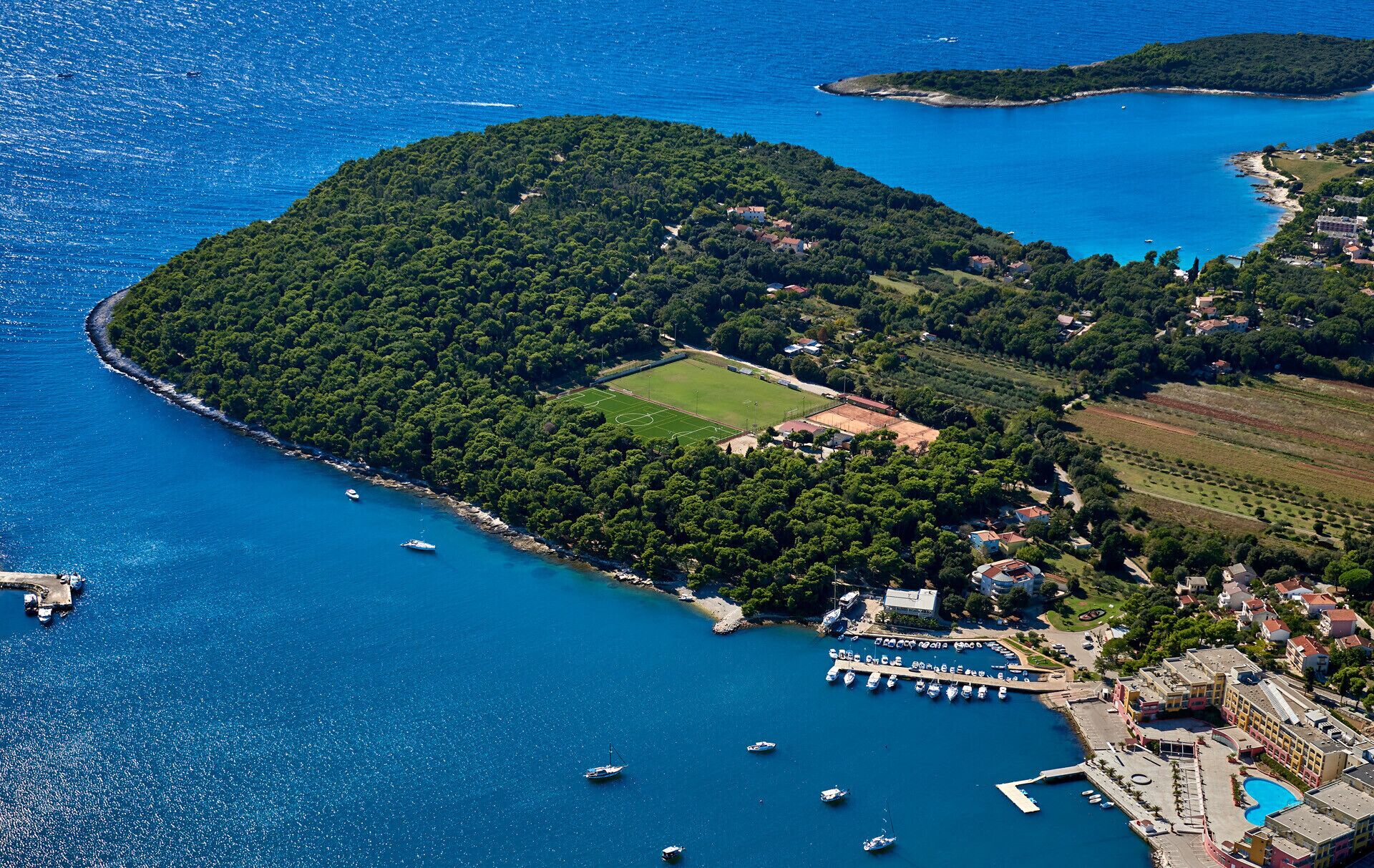 Vue aérienne d’une zone côtière comprenant un port de plaisance, un terrain de sport et des immeubles résidentiels.