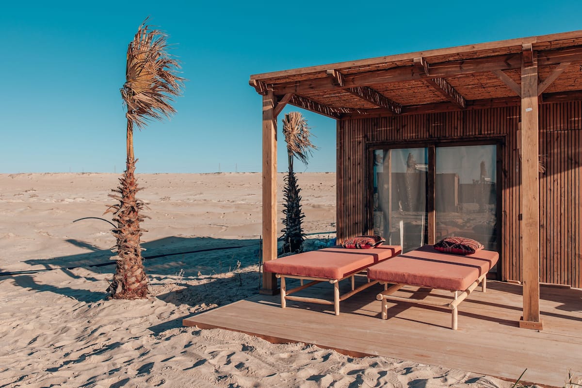 A wooden structure with two sunbeds and a glass door, set against a desert backdrop.