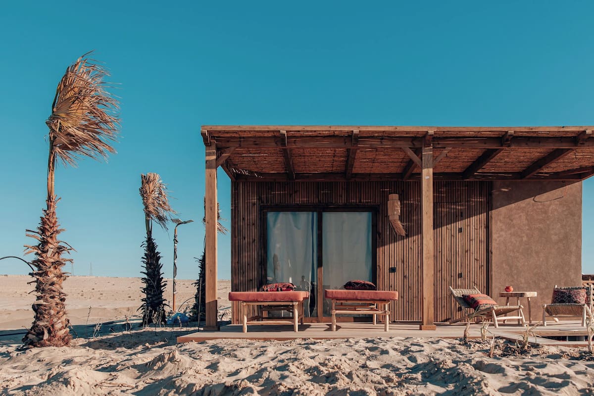 A wooden beach hut with two cushioned seating areas and a small table, surrounded by sand and palm trees.