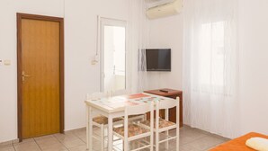A room with a wooden door, a white table with chairs, a TV mounted on the wall, and a bed with an orange blanket. - Studio apartment (Pirovac)