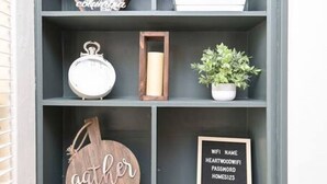 A grey shelving unit with various decorative items including a wooden sign with "gather," a framed WiFi password sign, and a small potted plant.