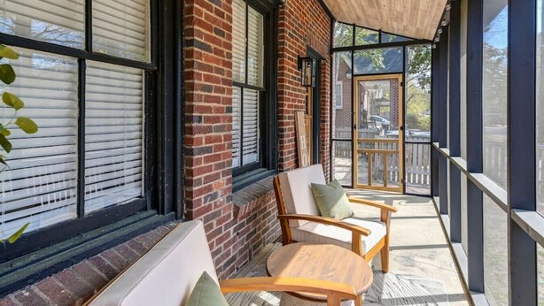 A covered porch with two wooden chairs and a bench, brick walls, large windows with blinds, and a view of the street.