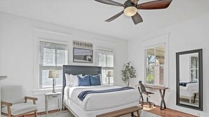 A bedroom with a bed, a desk, a chair, a ceiling fan, and a large mirror.