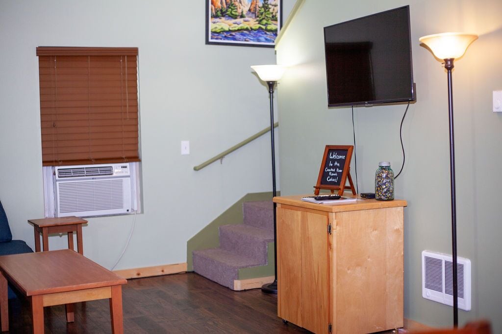 A room with a wooden desk, a small table, a staircase, a TV mounted on the wall, and a lamp.