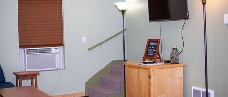 A room with a wooden desk, a small table, a staircase, a TV mounted on the wall, and a lamp.