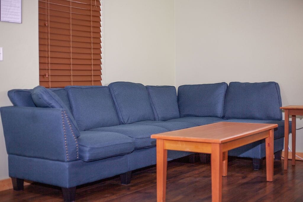 Un sofá seccional azul con una mesa de centro de madera en una habitación con piso de madera y una ventana con persianas.
