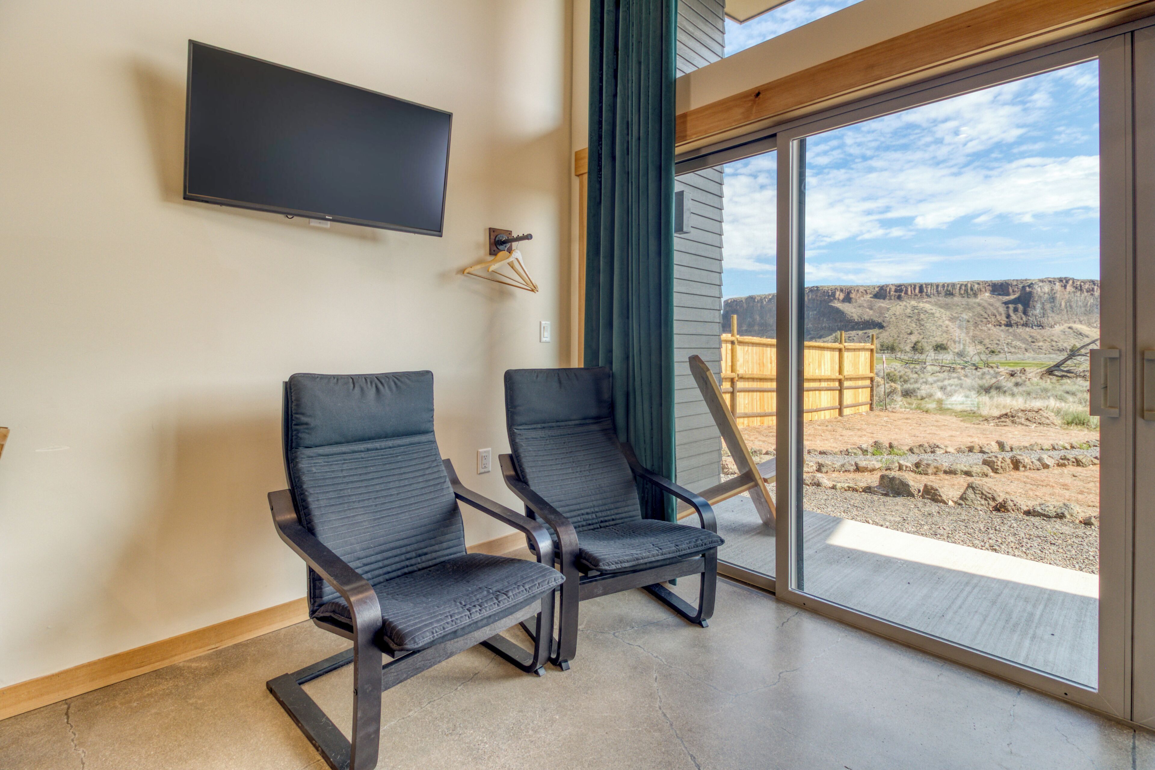 A room with two black chairs, a mounted TV, and a large sliding glass door leading to an outdoor area with a view of a mountainous landscape.