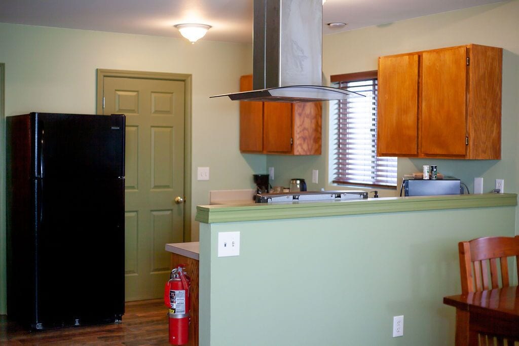 A kitchen with wooden cabinets, a black refrigerator, a fire extinguisher, and a window with blinds.