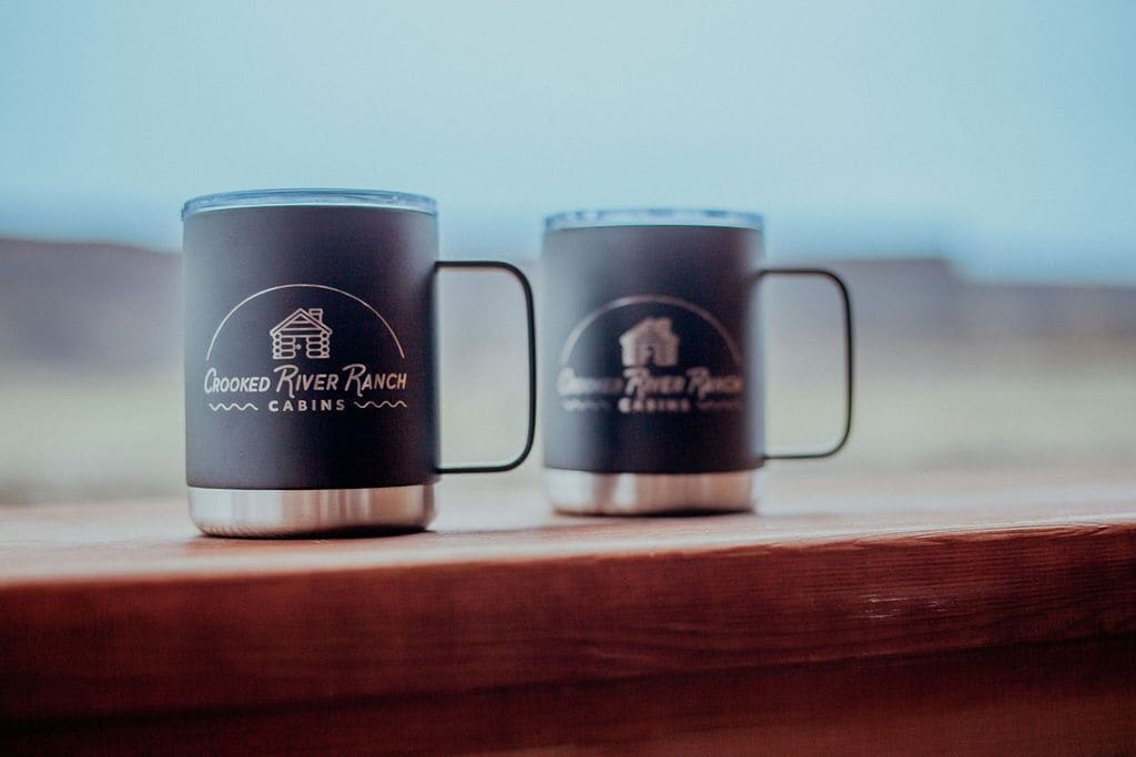 Two black mugs with handles, each labeled "Crooked River Ranch Cabins."