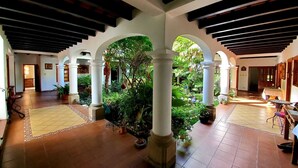 A spacious courtyard with tiled flooring, white columns, and arches, surrounded by lush greenery and potted plants.