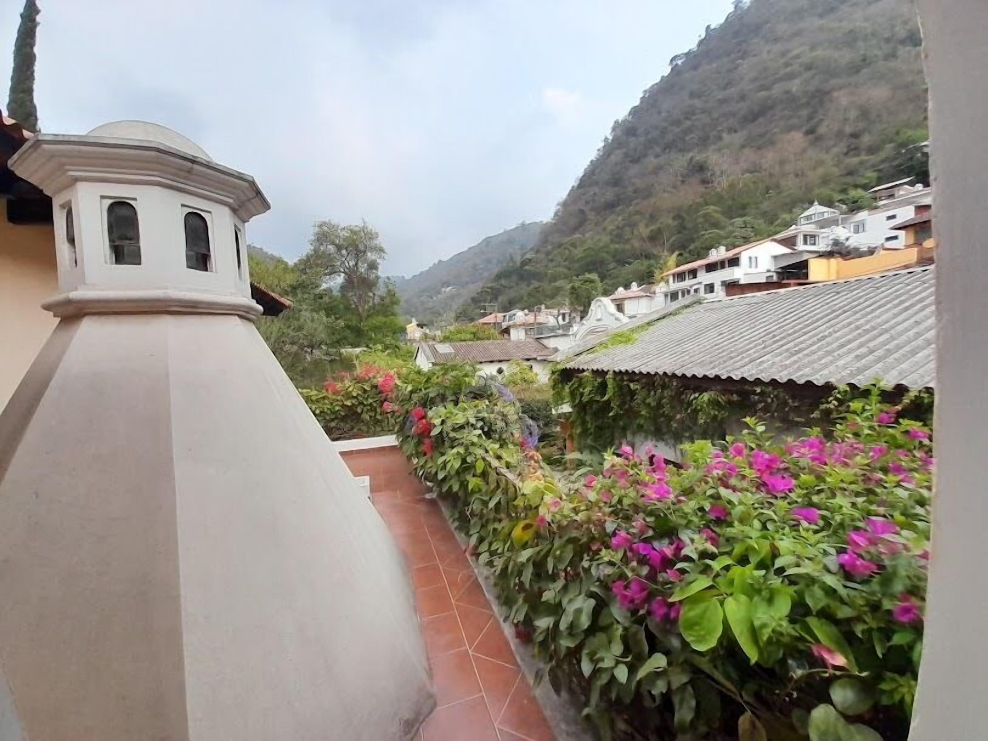 A residential area with a white building featuring a small tower-like structure, surrounded by lush greenery and pink flowers.