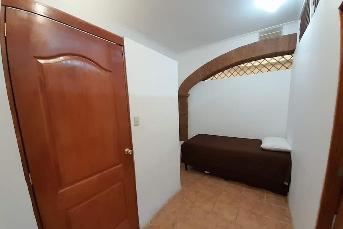 A small room with a wooden door, a bed with a mattress and pillow, and a tiled floor.