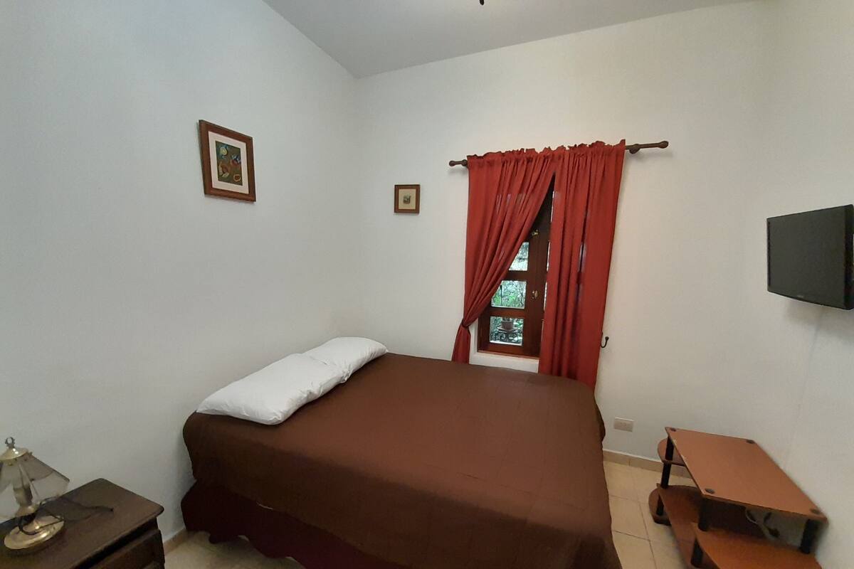 A bedroom with a single bed, a small table, a chair, a TV, and a window with curtains.