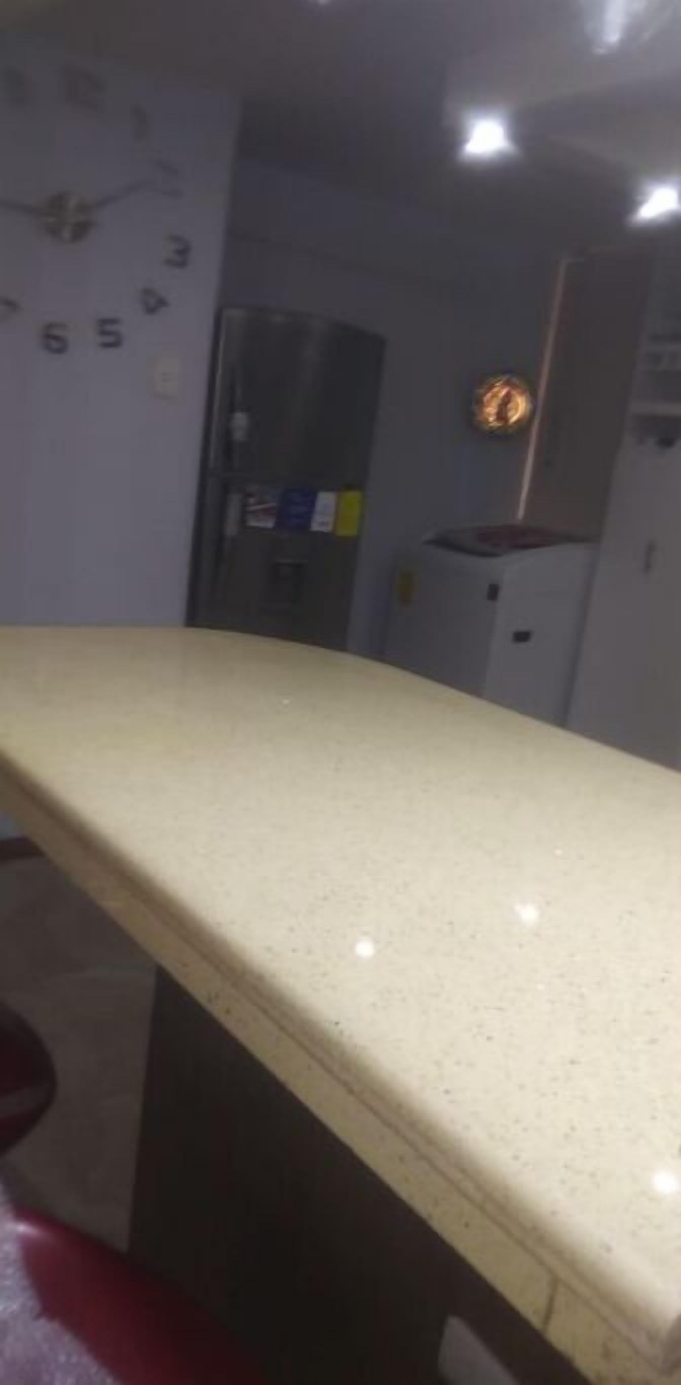 A room with a long, beige countertop, a clock on the wall, and a refrigerator in the background.