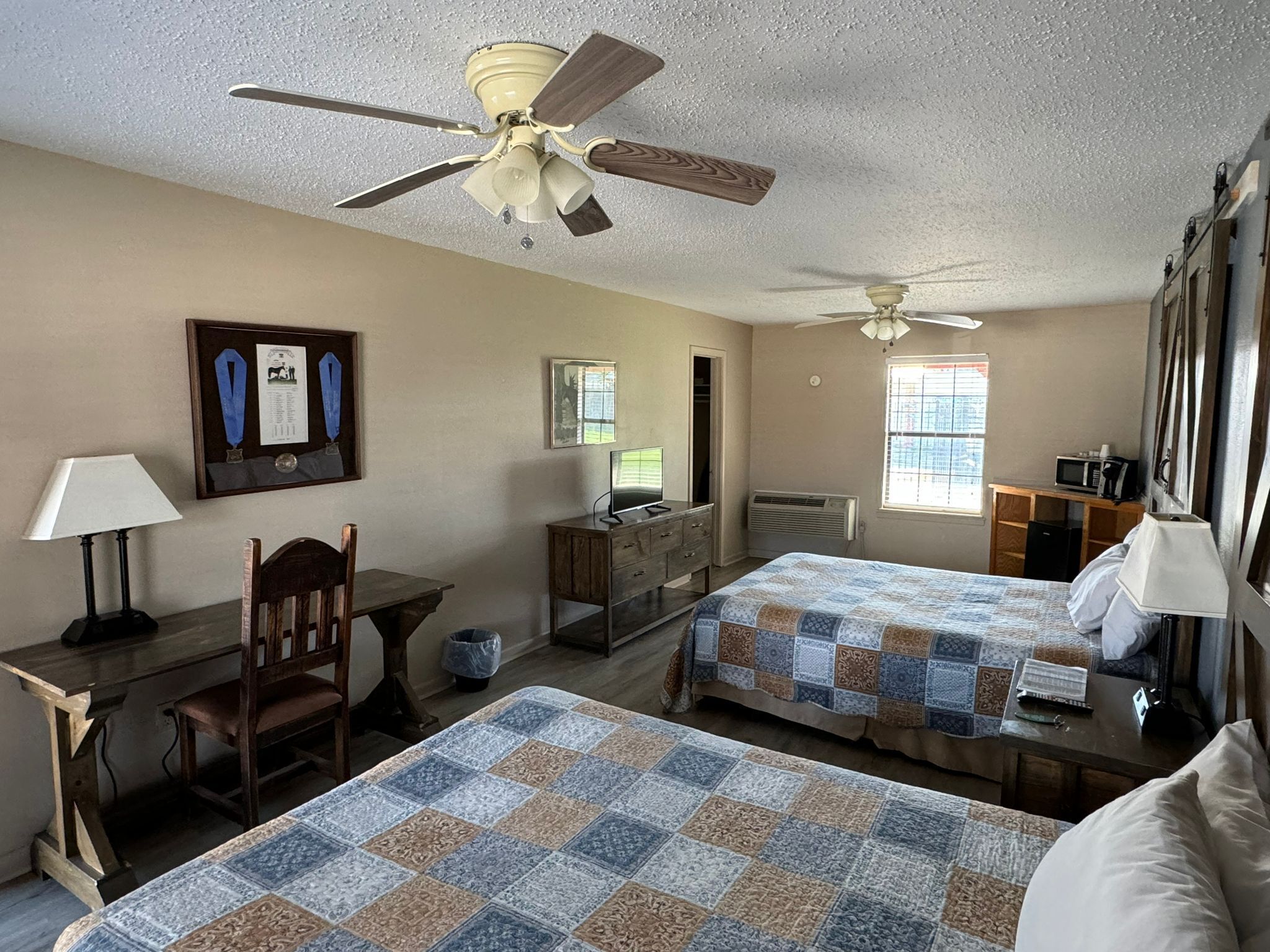 A hotel room with two beds, a desk, a chair, a TV, and a ceiling fan.