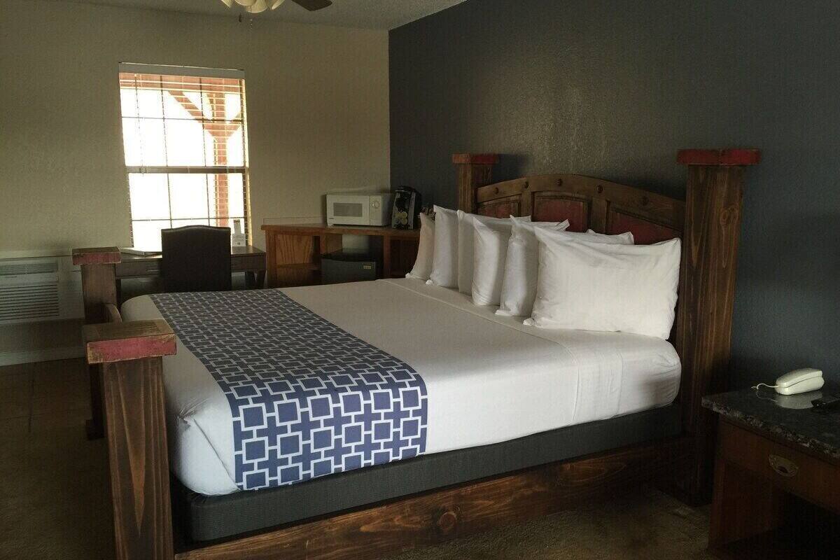 A bedroom with a wooden bed, a desk, a chair, a microwave, and a window with blinds.