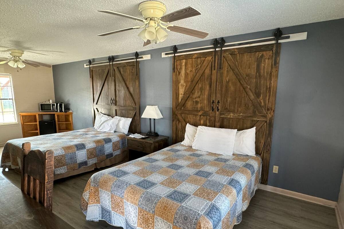 A bedroom with two beds, wooden headboards, a ceiling fan, a nightstand with a lamp, and a small desk with a microwave.