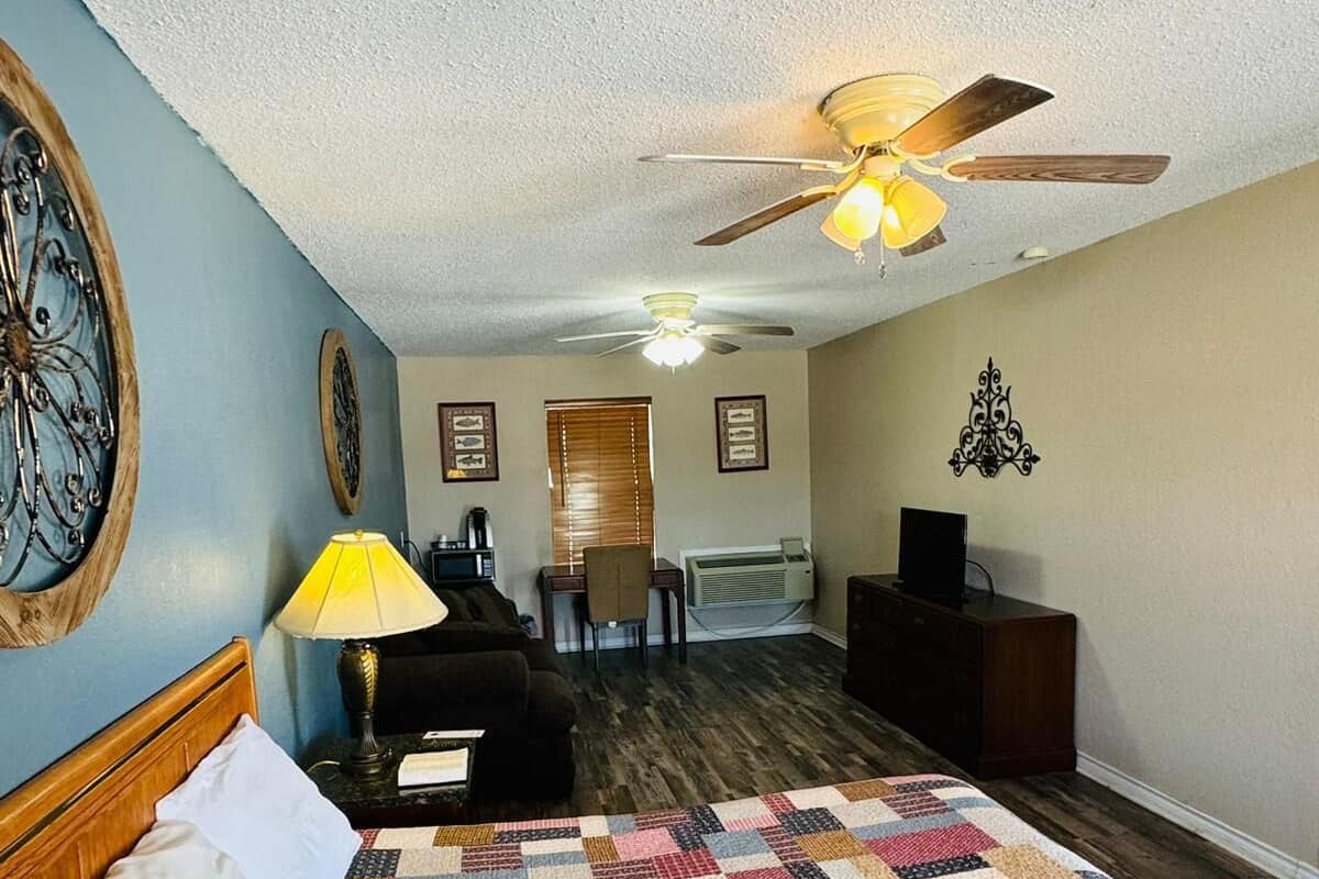 A hotel room with a large bed, a ceiling fan, a desk with a lamp, and a view of the hallway.
