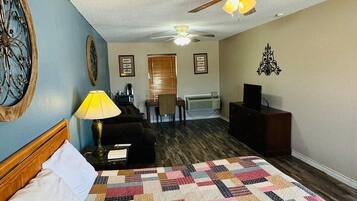 Quarto de hotel com uma cama grande, um ventilador de teto, uma mesa com abajur e vista para o corredor.