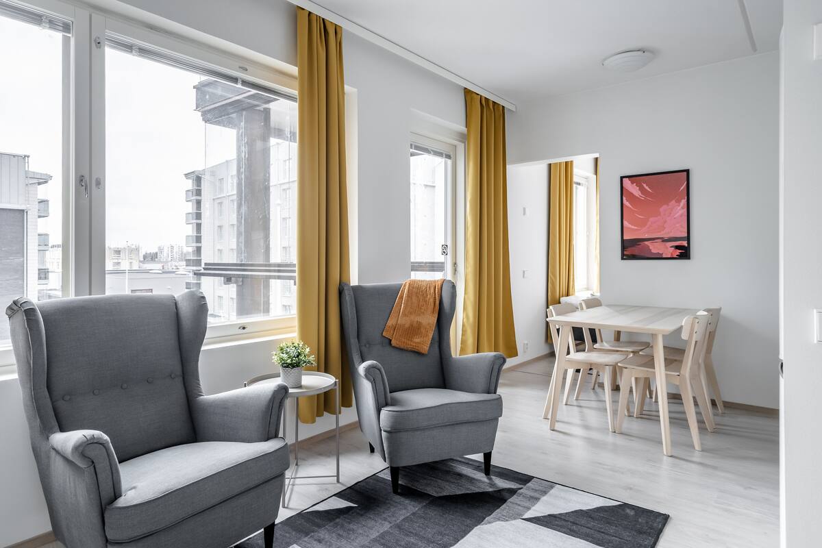 A modern living room with a grey armchair, a small table with a plant, and a dining area with a white table and wooden chairs.