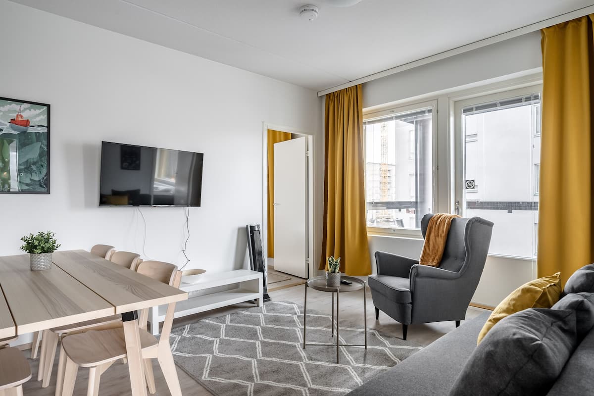 A modern living room with a wooden dining table, grey sofa, and a TV mounted on the wall.