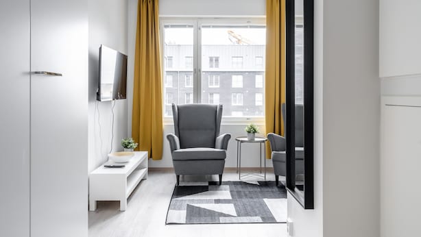 Un salon moderne avec un fauteuil gris, une petite table d’appoint ornée d’une plante et une grande fenêtre avec des rideaux jaunes.