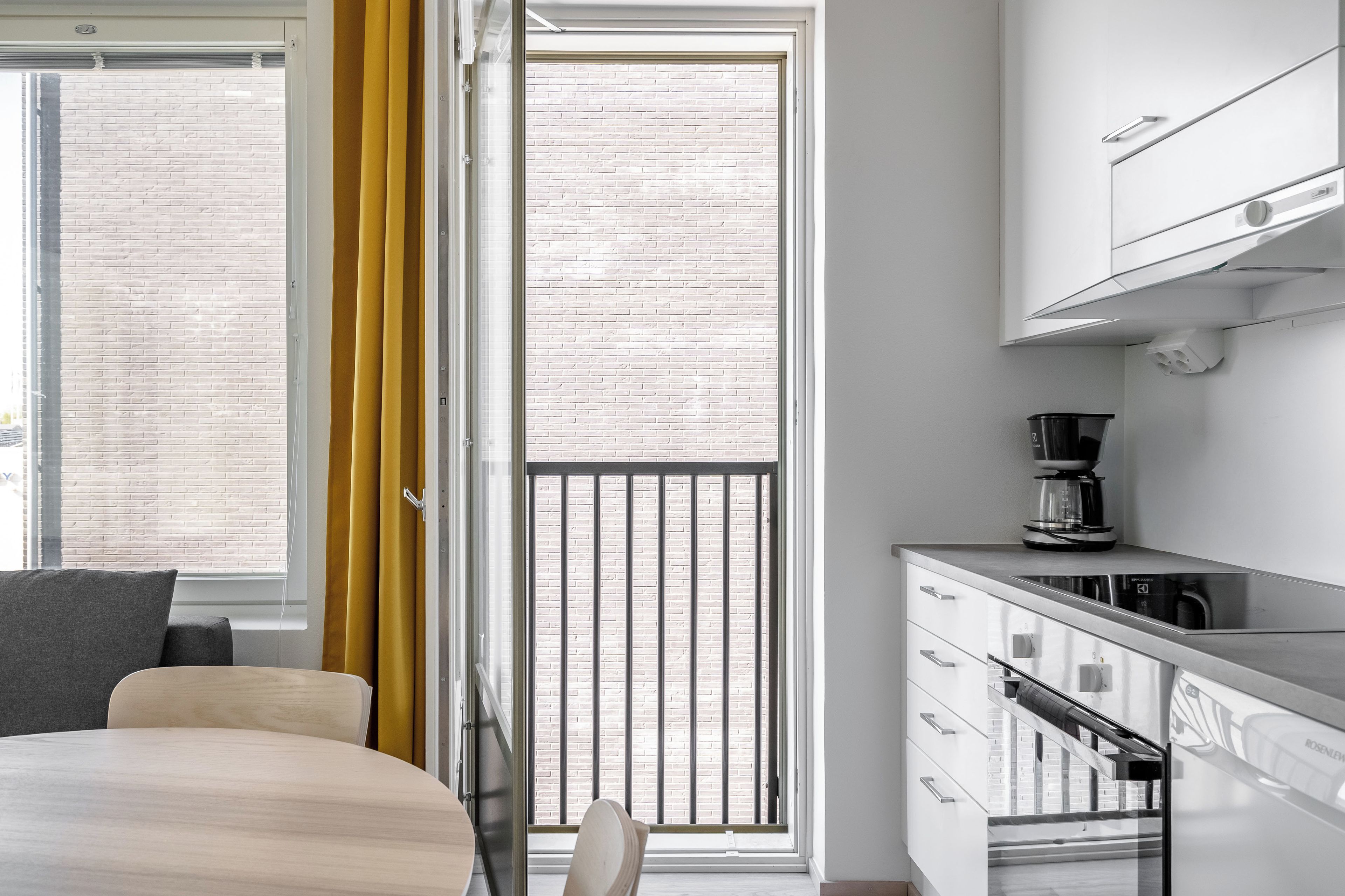 A modern kitchen with a coffee maker, a dining area with a bed, and a view of a brick building through a window.