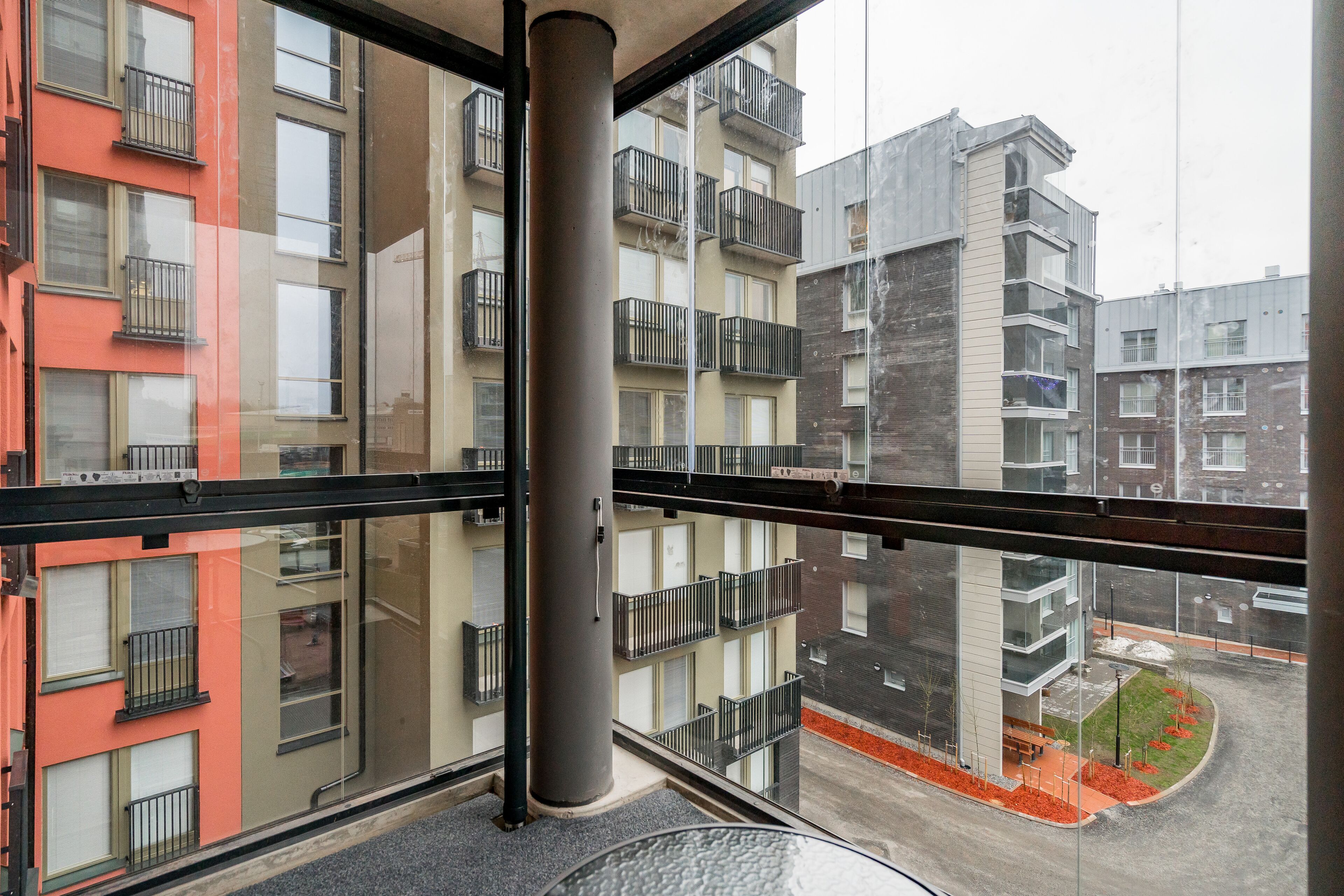 A multi-story residential building with balconies, a glass facade, and a visible courtyard with a bench and small trees.
