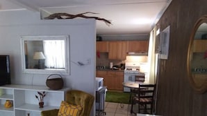 Un salon comprenant un canapé, un téléviseur et un coin repas avec une table et des chaises. Au fond, on aperçoit une cuisine avec des meubles en bois et un four à micro-ondes.