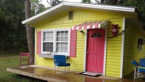 Une petite maison jaune avec une porte rouge et des volets roses, une véranda avec des chaises bleues et une terrasse en bois.