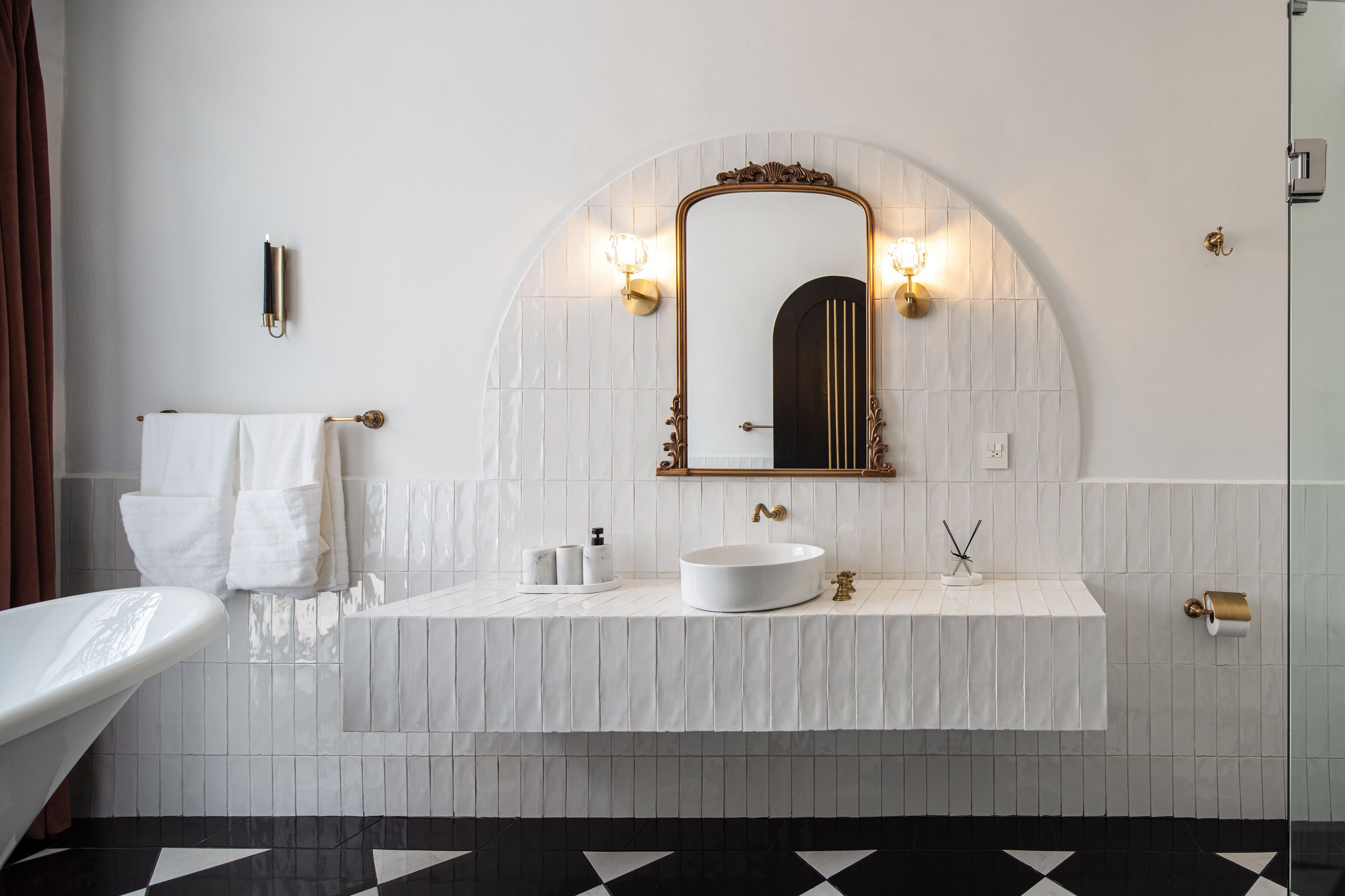 A modern bathroom with a white vanity, a round sink, and a large mirror with an arched frame. There are two wall-mounted lights above the mirror and a towel rack with white towels.